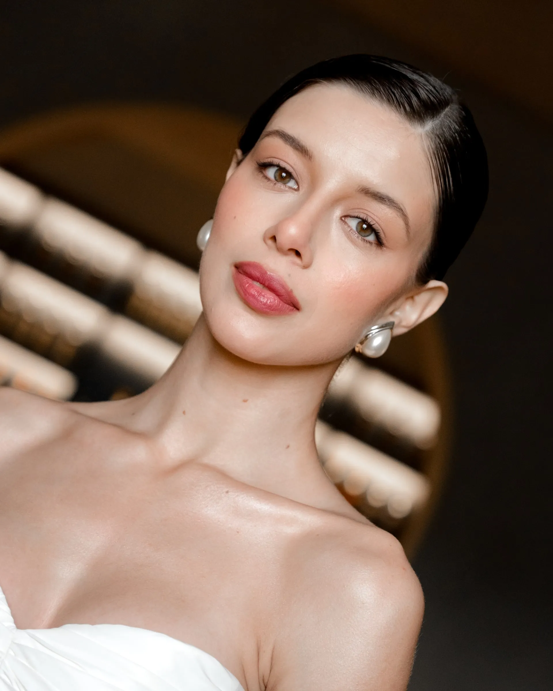 Beauty close-up with luminous skin, a clean sculpted brow and a glossy rose lip against slicked-back hair.