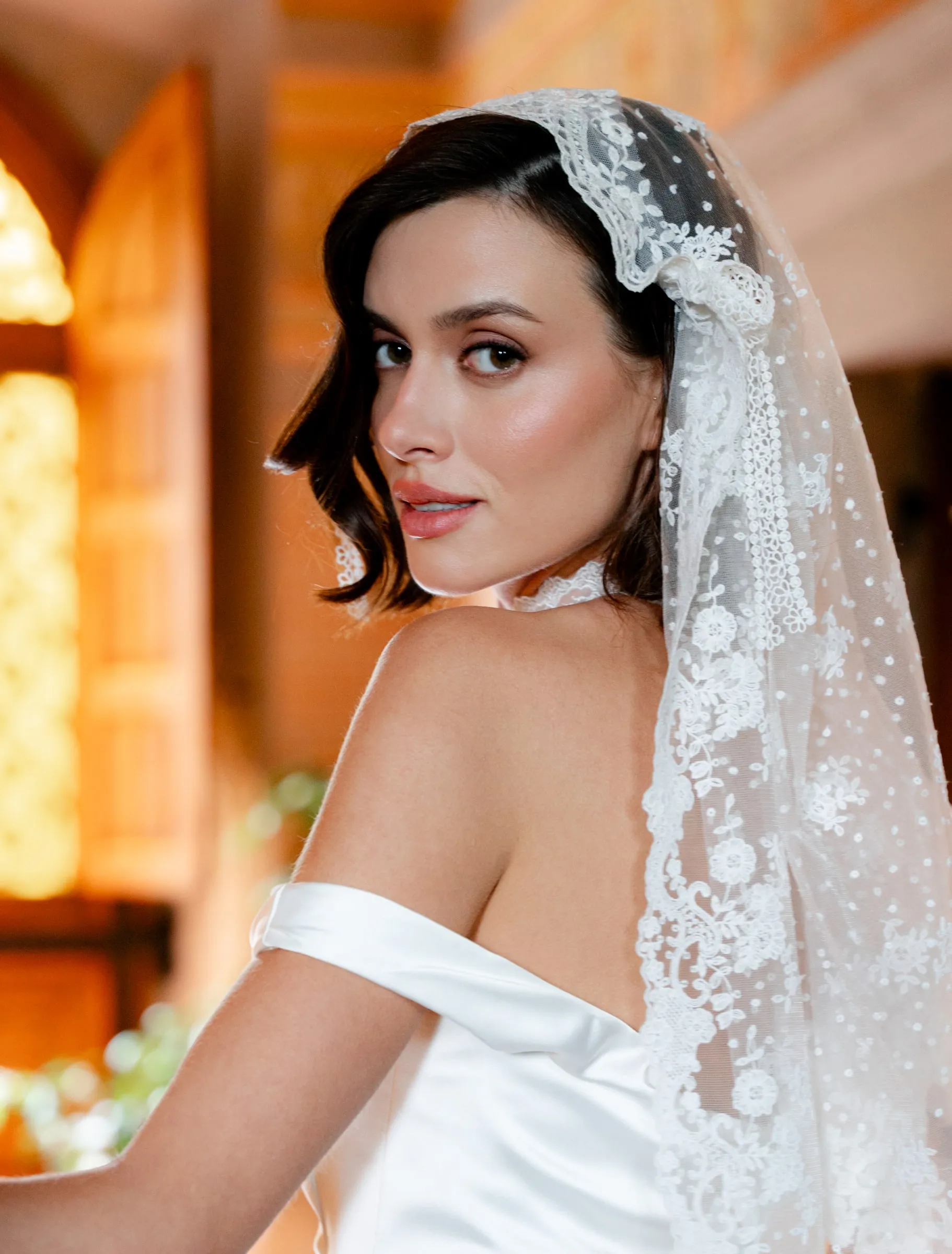 Bride in a lace mantilla veil with a dewy base, softly smoked liner and a muted berry lip in warm light.