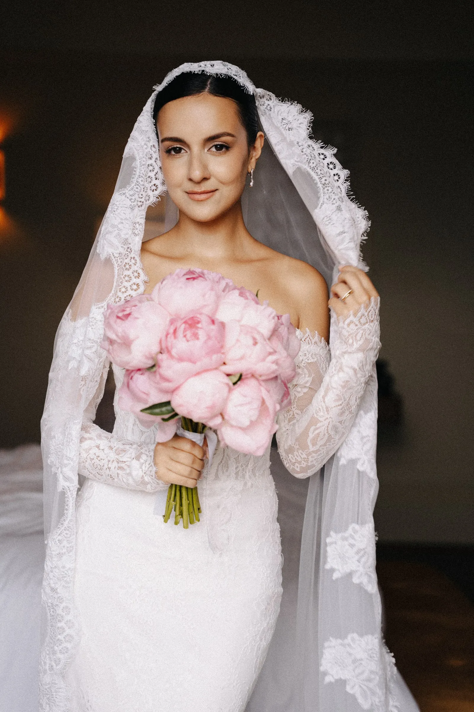 Bride under a lace veil holding pink peonies, with soft-focus skin, a warm eye and a rosy lip.