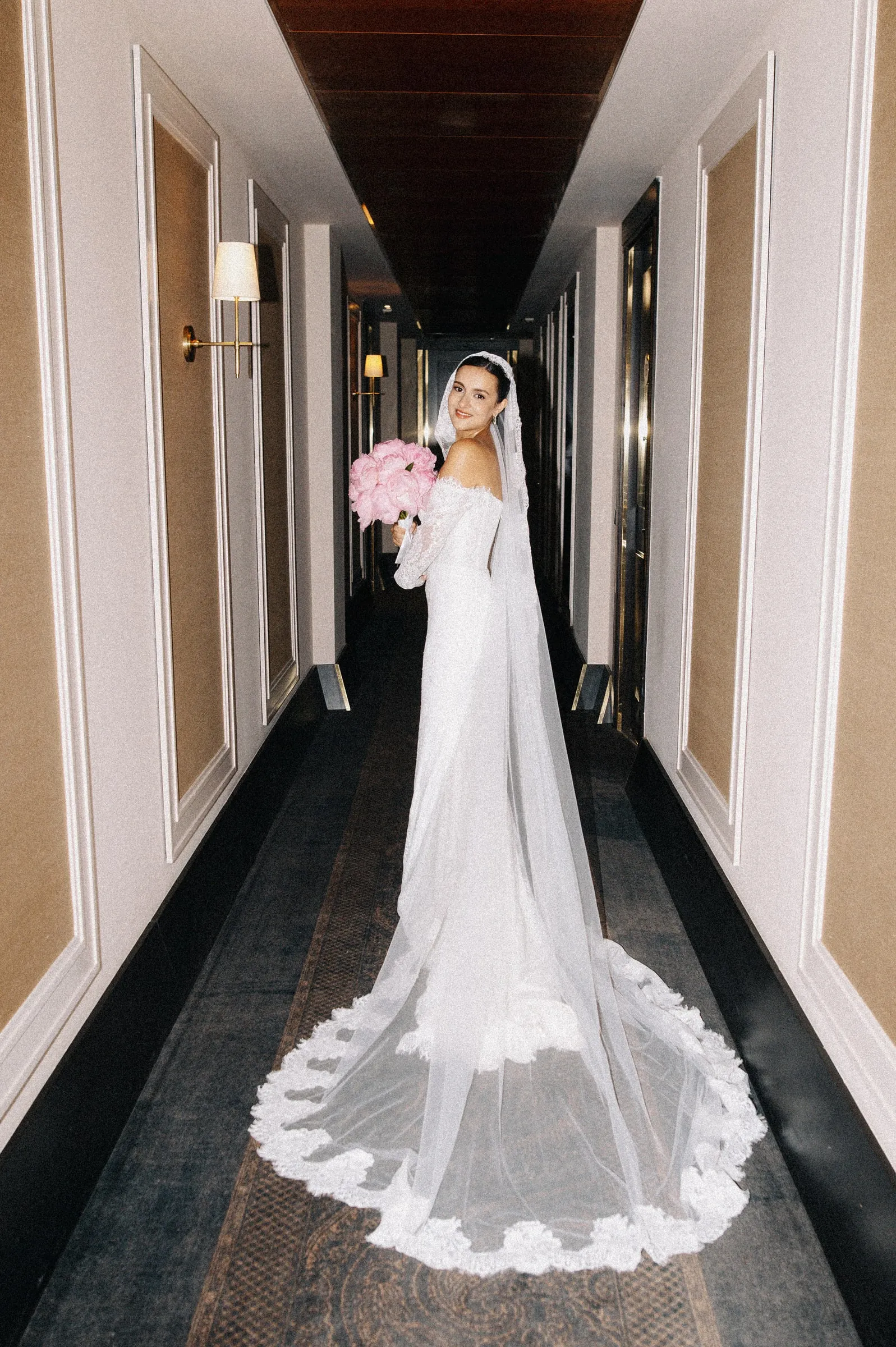 Bride photographed down a hotel corridor with a long lace-edged veil, wearing a clean long-wear base.