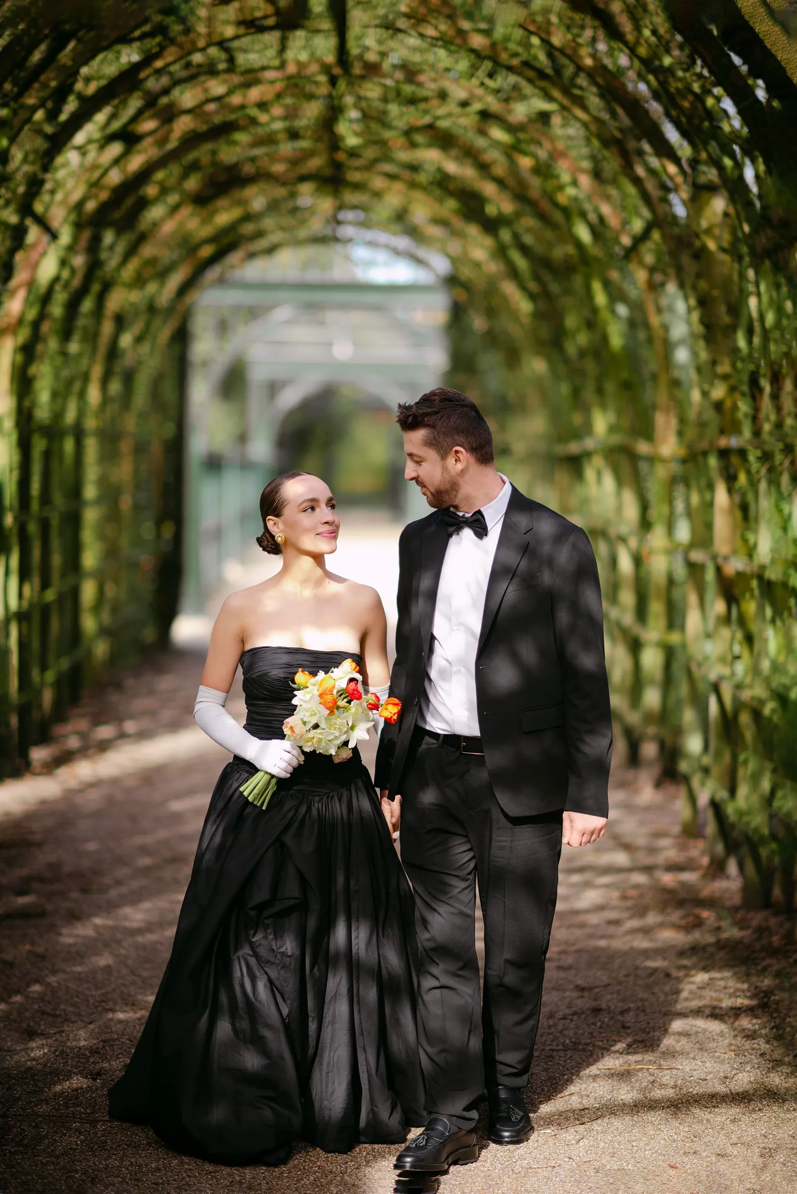 Couple walking beneath a green garden arch, the bride in a black gown with a defined eye and pale lip.